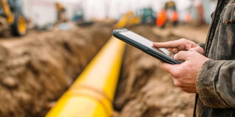 Construction worker using a tablet to monitor progress on a site with heavy machinery and a large yellow pipe being laid in a trench