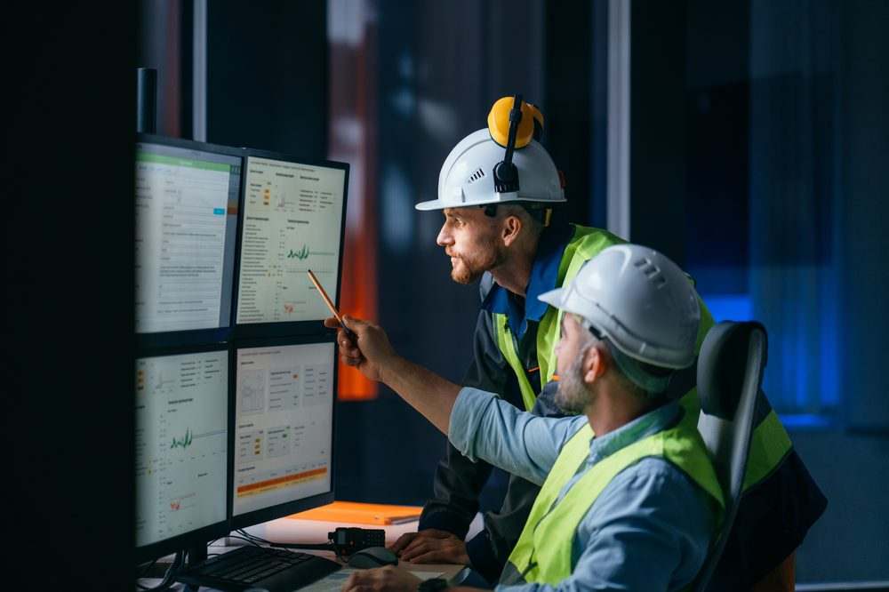 Two engineers in hard hats reviewing operational dashboards on multiple monitors
